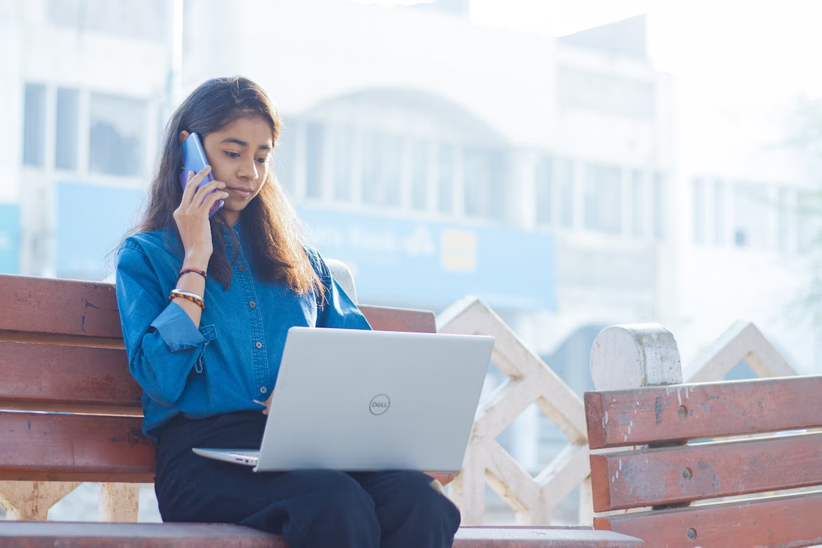 Woman sitting on bench, looking at laptop and using phone