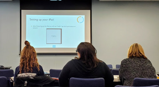 Three women sitting in chairs looking at presentation on white board